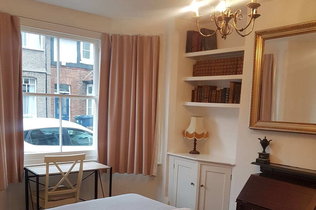 A hotel room with a large bed, a desk, a chair, a bookshelf, a dresser, and a window with curtains.