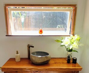 A bathroom with a wooden vanity, a stone basin, a bottle, and a vase with flowers. - Charming beach  Cottage (Bay View)