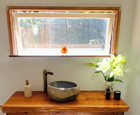 A bathroom with a wooden vanity, a stone basin, a bottle, and a vase with flowers.