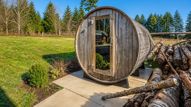A wooden barrel structure with a door, logs, and a grassy area with trees.
