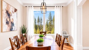 A dining room with a wooden table, chairs, a hanging light fixture, a window with a forest view, and a painting on the wall.