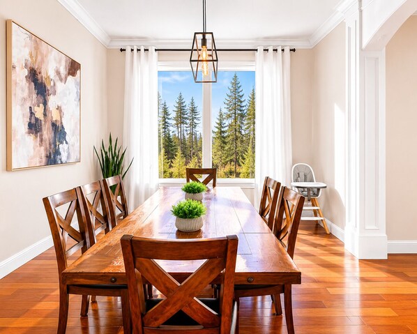 A dining room with a wooden table, chairs, a hanging light fixture, a window with a forest view, and a painting on the wall.