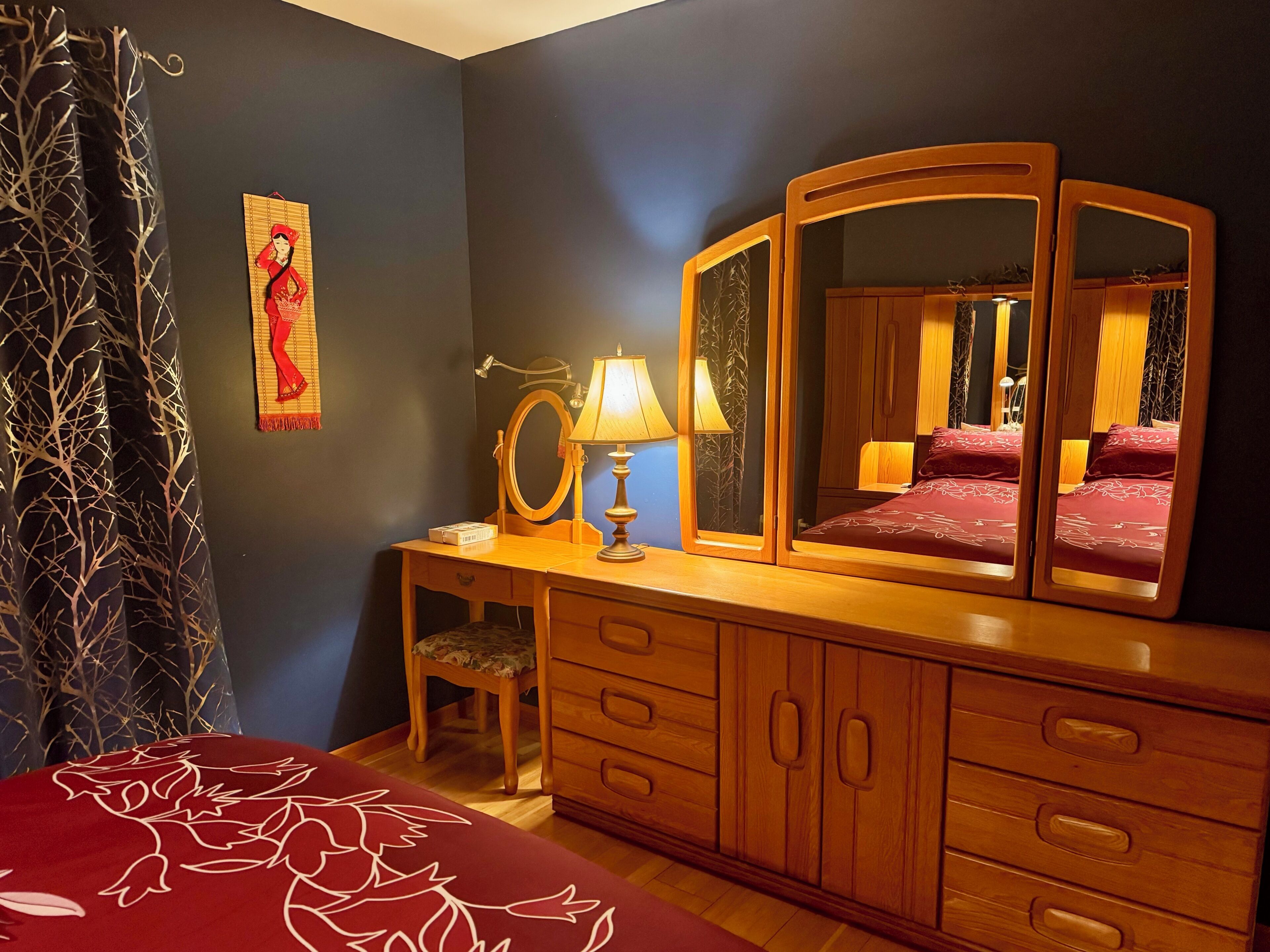 Un dormitorio con cómoda de madera, espejo, mesita de noche con lámpara, cama con colcha floral roja y un cuadro colgado en la pared.
