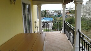 A balcony with a wooden table, a white railing, and a view of a residential area with houses and a blue-roofed building.
