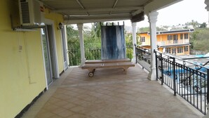 A covered balcony with a wooden bench, a black curtain, and a view of a yellow building and palm trees.