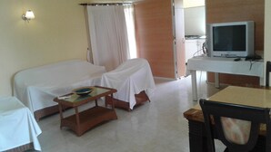 A room with a bed covered in white, a small table with a bowl, a television on a stand, and a desk with a computer.