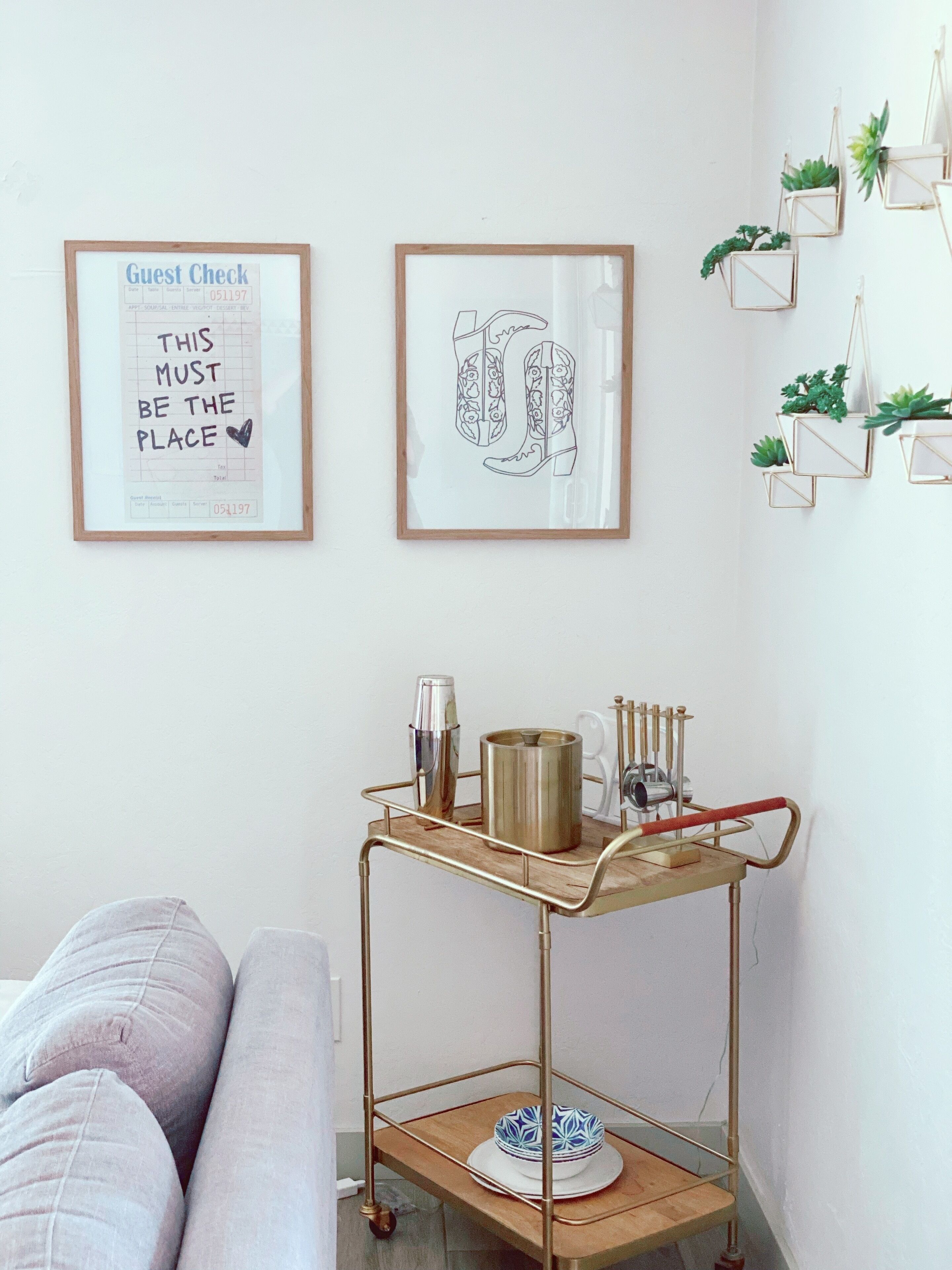 A room with a minimalist decor, featuring a framed 'Guest Check' sign, a decorative shoe illustration, and a metal cart with a bowl and candle holder.