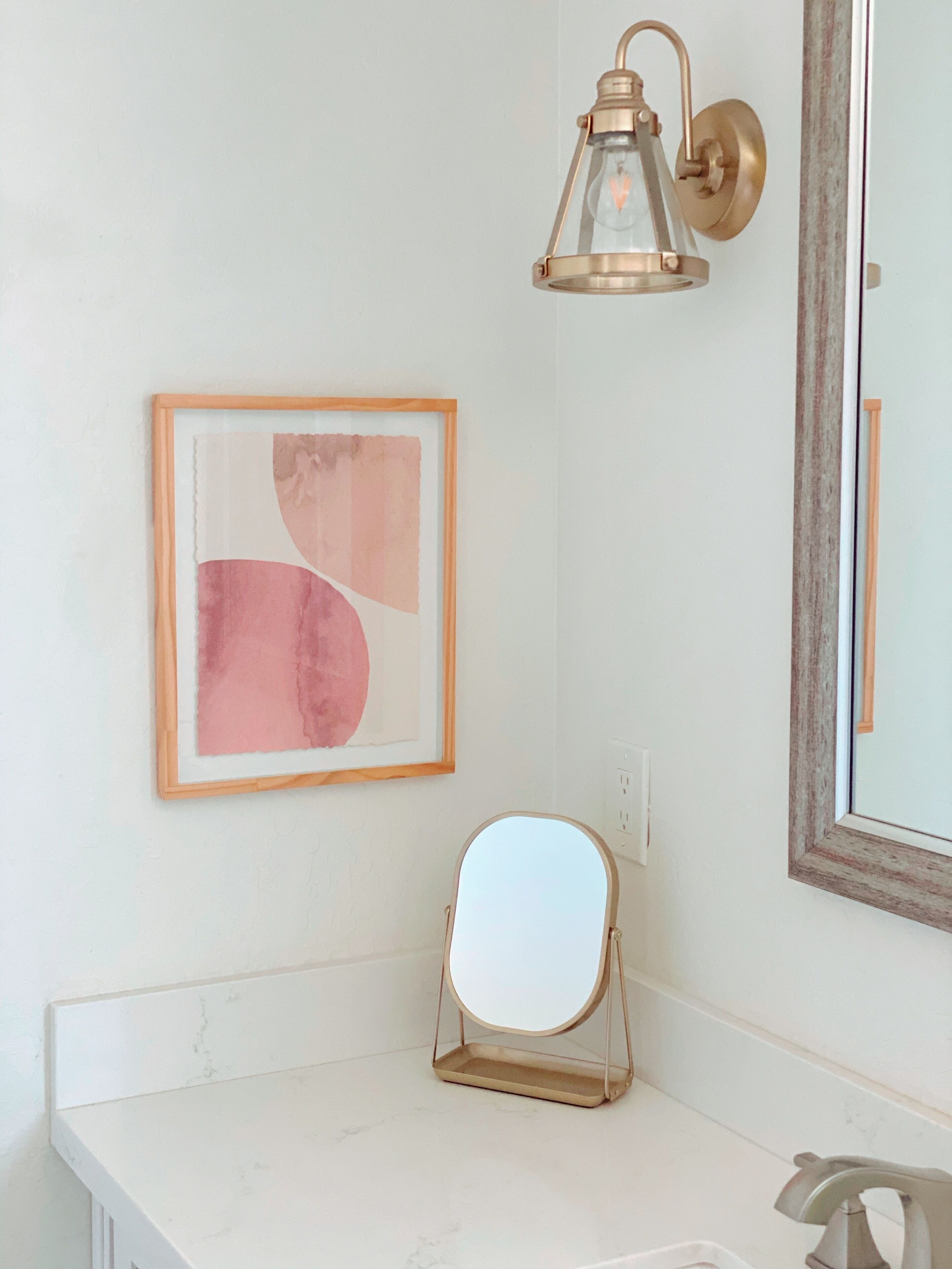 A bathroom with a white countertop, a framed abstract painting, a wall-mounted light fixture, and a mirror with a gold frame.