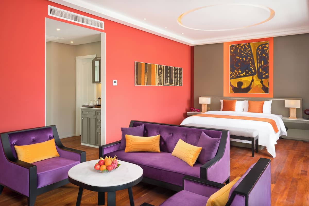 A hotel room with a bed, purple sofas, a small table with fruit, a painting, and a bathroom visible through an open door.