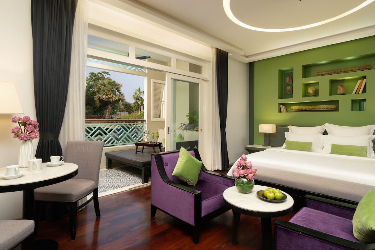 A hotel room with a bed, a seating area with a table and chairs, a balcony with a view of palm trees, and a green accent wall with framed pictures.