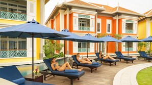 Un área junto a la piscina con sillones de descanso y sombrillas, una piscina y un edificio con fachada amarilla y detalles decorativos verdes.