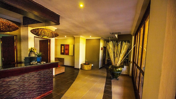 A hotel lobby with a reception desk, a seating area, a hallway with a plant, and a wall with framed pictures. - Coyotes Hotel and Conference Centre (Mbombela)