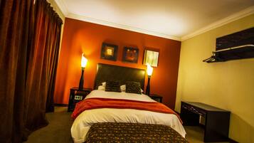 Quarto de hotel com uma cama grande, duas mesas de cabeceira, um criado-mudo, um banco e uma prateleira fixada na parede.