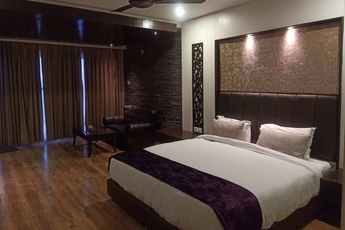 A modern hotel room with a large bed, a ceiling fan, a dark leather armchair, and a view of the outdoors through a large window.
