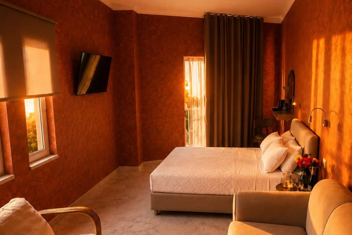 Quarto de hotel com cama, sofá, poltrona, ventoinha no teto, televisão e janela com cortinas.