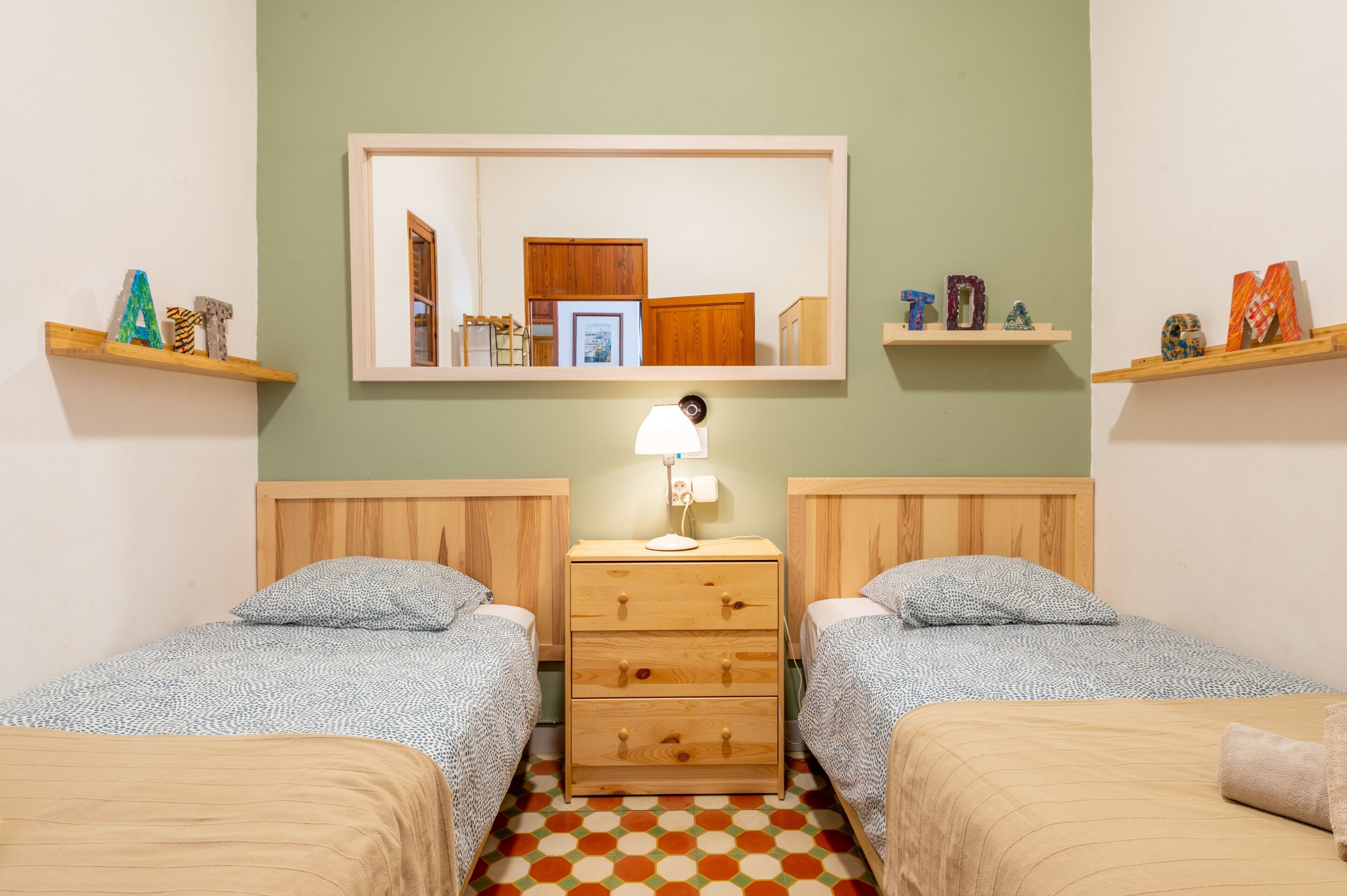 Una habitación con dos camas, una mesita de noche, un espejo y repisas con objetos decorativos.