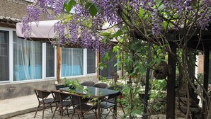 En terrasse med et bord og stole, en lilla blomsterplante, der hænger ned, og et vindue med et gennemsigtigt gardin.