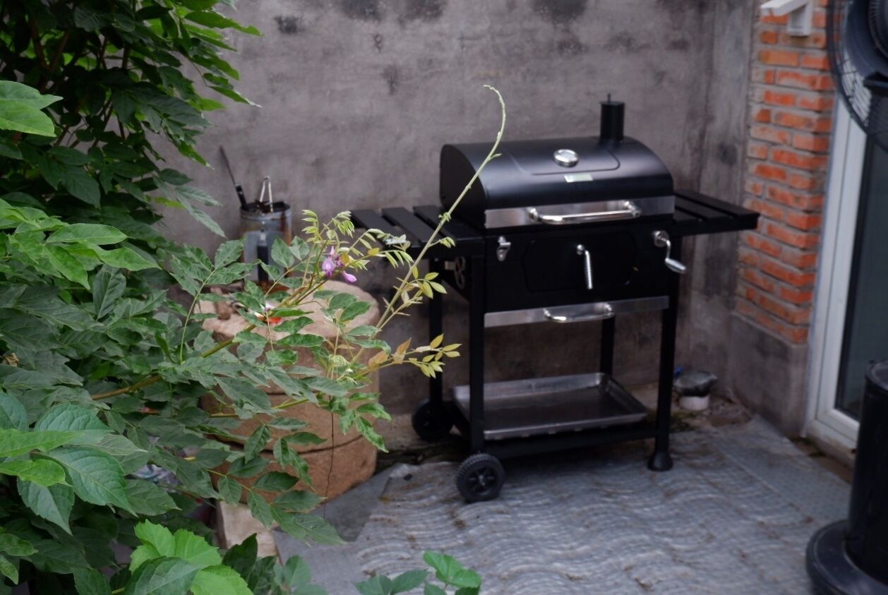 A portable grill with a cart, a potted plant, and a brick wall in the background.
