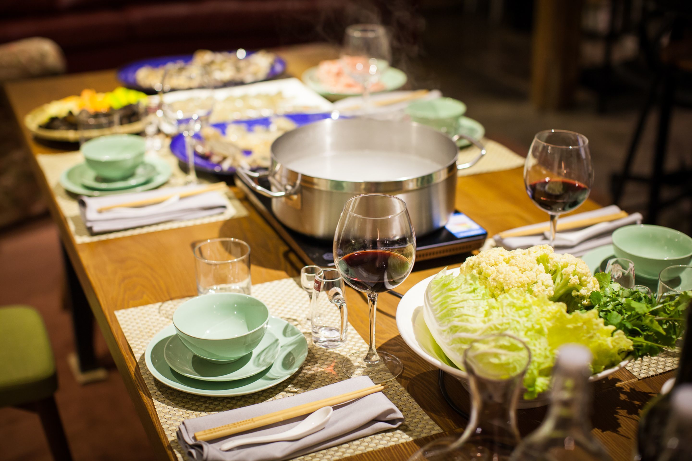 A dining table set with various dishes, a pot of boiling water, and glasses of red wine.