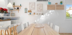 A modern kitchen with white cabinetry, a wooden dining table, and chairs. There are wall-mounted shelves with decorative items and a window providing natural light.