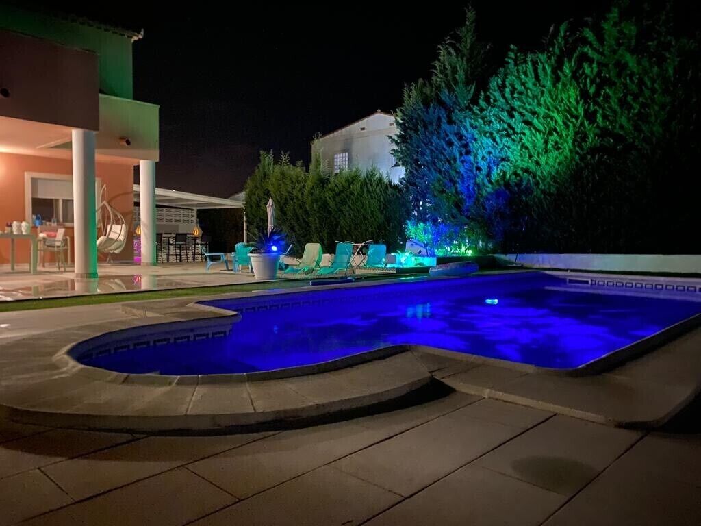 A swimming pool with blue lighting, surrounded by lounge chairs and a building with a covered patio.