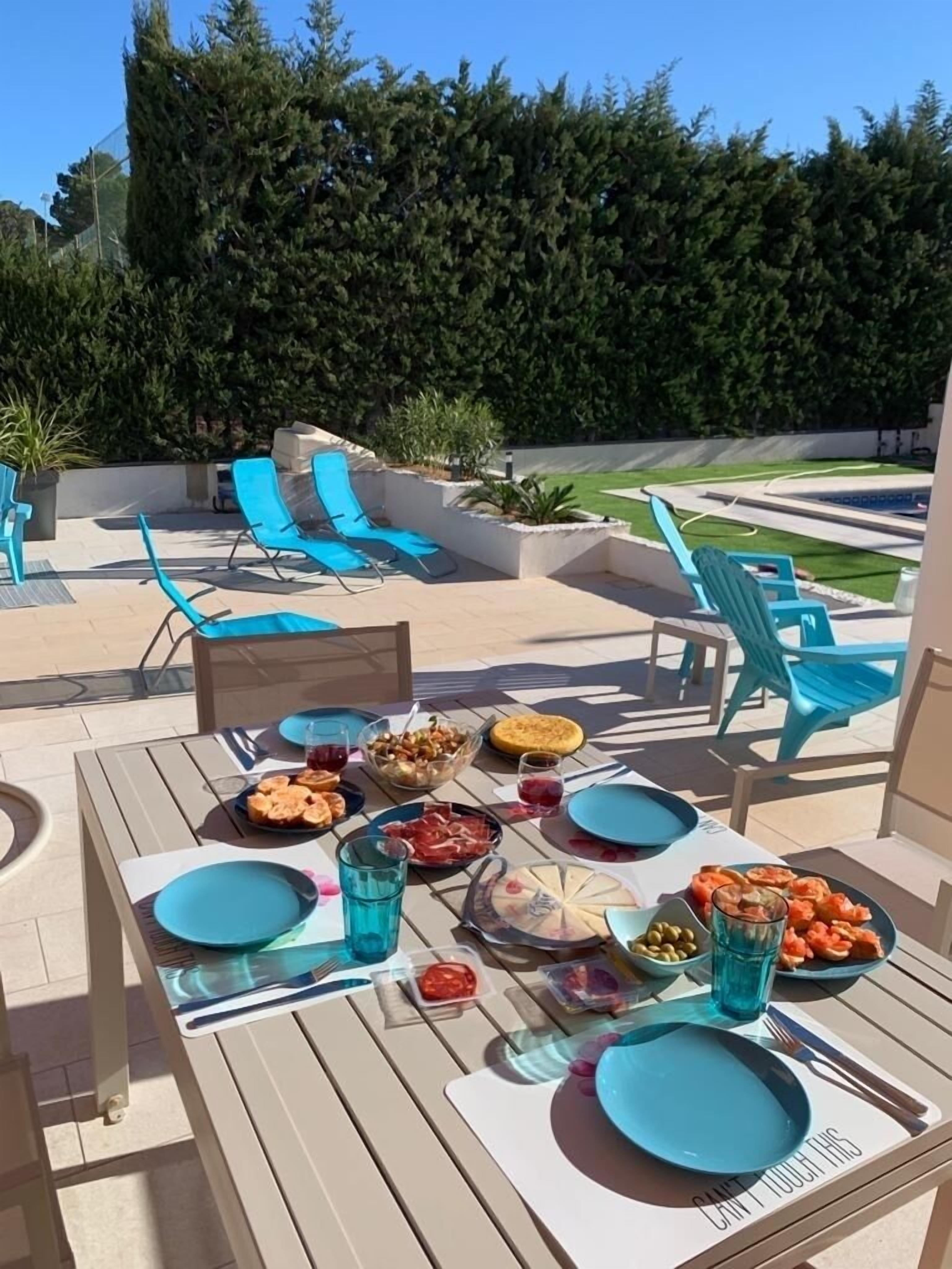 An outdoor dining area with a table set for a meal, surrounded by blue chairs, and a pool in the background.