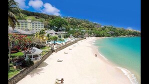 A beach with a resort building, clear turquoise water, and a sandy shore. - Apartment with a million dollar view and a wrap-around balcony with beach access (Castries)