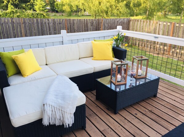 En terrasse med en hvid sofa, gule puder, et hvidt pled og to lanterner på et bord med glasplade.
