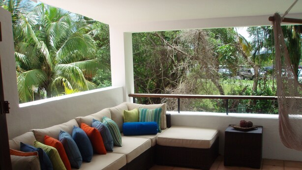 A sunlit room with a beige sofa, colorful cushions, a small table with fruit, and a view of lush greenery outside.