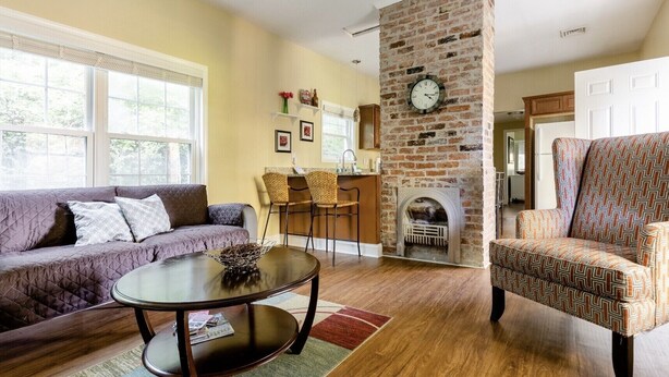 A living room with a brick fireplace, a sofa, a patterned armchair, and a dining area with wicker chairs.