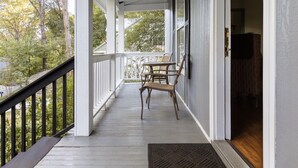 A covered porch with a wooden table and chairs, a black railing, and a view of trees and a house in the background.
