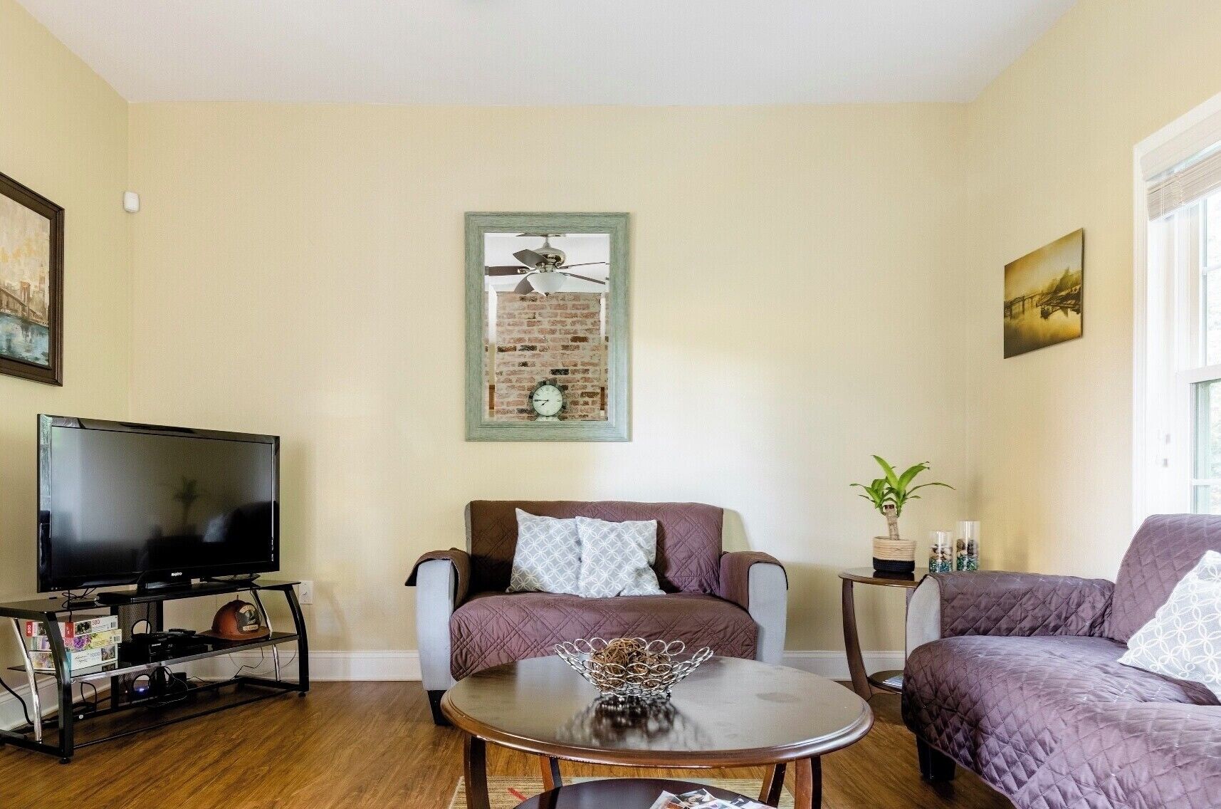 A living room with a flat-screen TV, a round coffee table, two sofas, a small side table with a plant, and a framed picture on the wall.