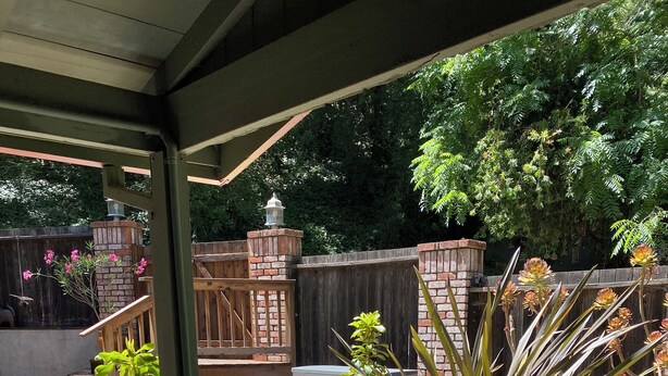 Una terraza de madera con mobiliario exterior, plantas en macetas y un muro de ladrillo al fondo.