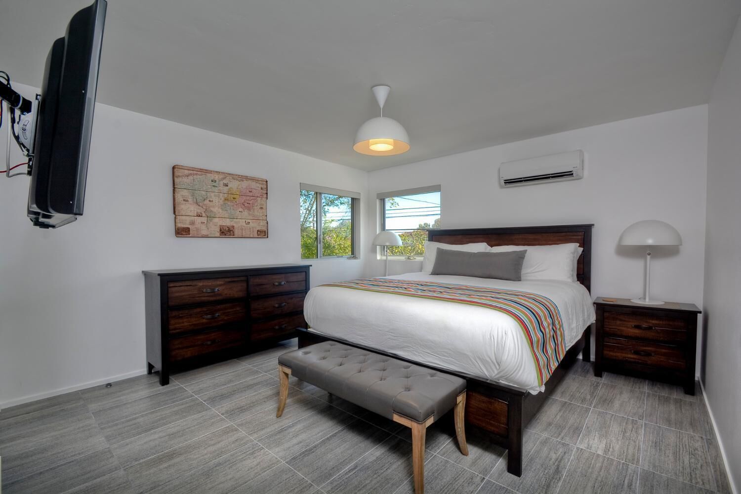 A bedroom with a large bed, a wooden dresser, a bench, a map on the wall, and a window with a view.