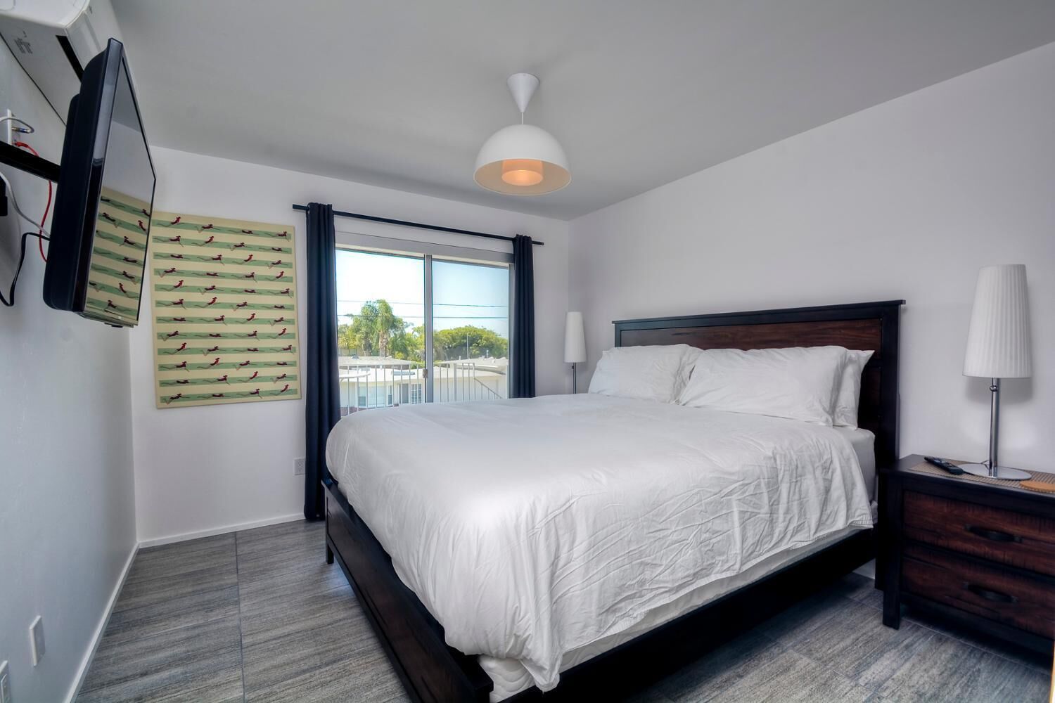 A modern hotel room with a large bed, a flat-screen TV mounted on the wall, a ceiling light, and a window with a view of the outside.