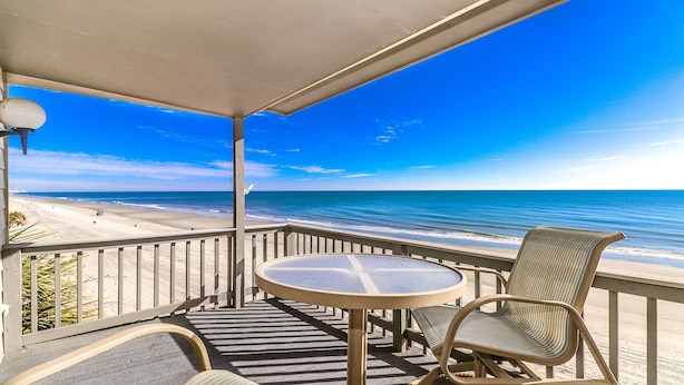 A balcony with a round table and two chairs, overlooking a beach with a clear blue sky and calm sea. - Beachfront/Pool Murrells/GardenCity/Surfside 
Wkly/MilitaryFirePoliceNurse Discs (Murrells Inlet)