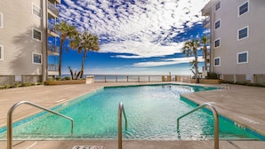 A swimming pool with a view of the ocean, surrounded by buildings with balconies and palm trees. - Beachfront/Pool Murrells/GardenCity/Surfside 
Wkly/MilitaryFirePoliceNurse Discs (Murrells Inlet)