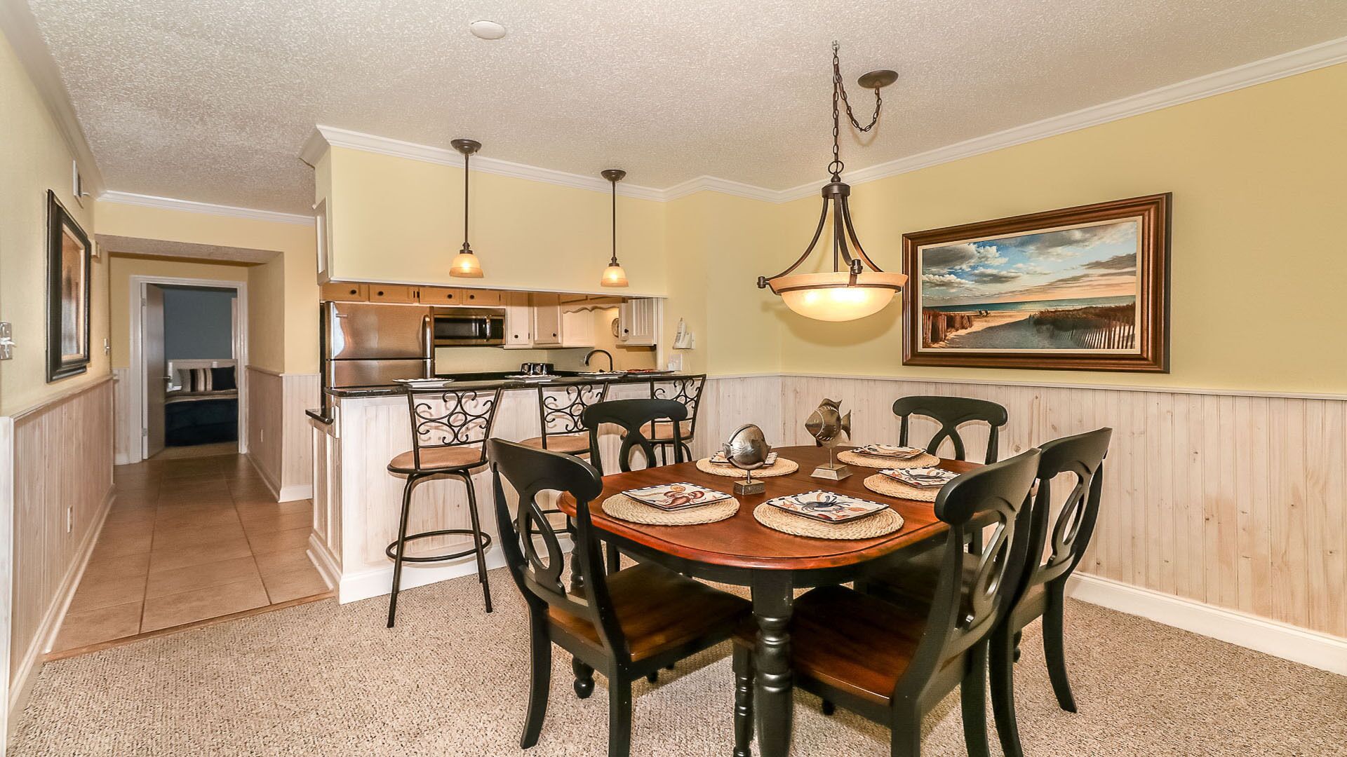 A dining area with a wooden table and chairs, a kitchen in the background, and a framed landscape painting on the wall.