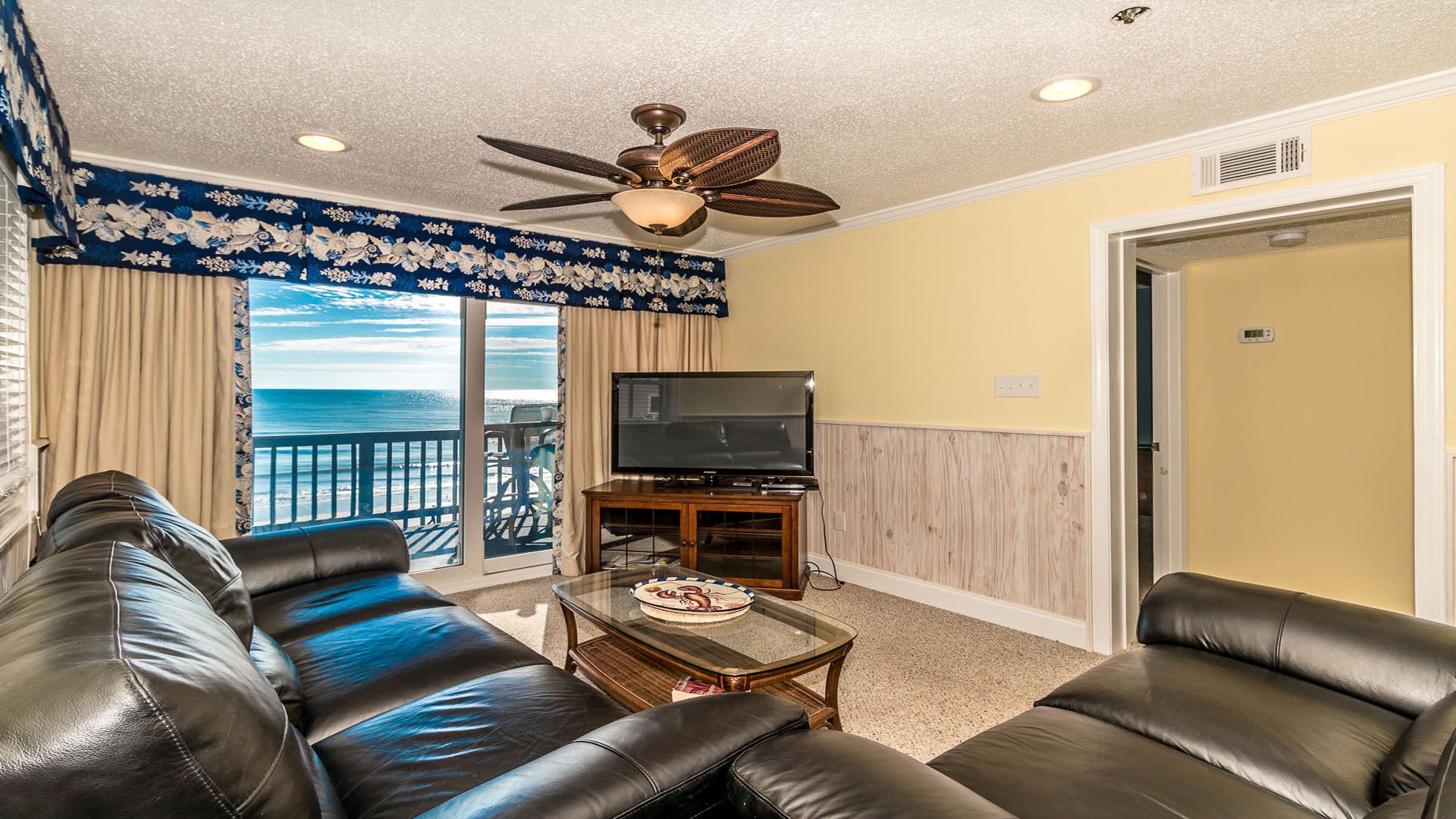 A living room with a balcony, leather sofas, a television, and a ceiling fan.