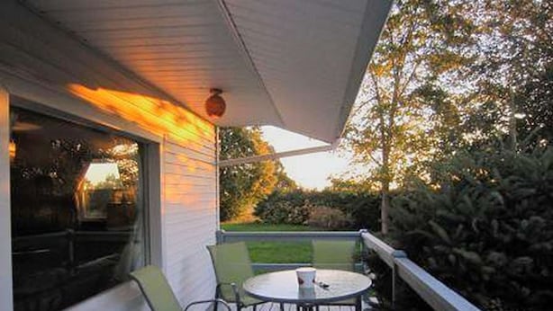 A covered outdoor seating area with a table and chairs, a window, and a view of the garden.