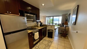 Cocina compacta con refrigerador, estufa, microondas y lavamanos. Hay un basurero pequeño y un tapete estampado. La cocina da a un comedor con mesa y sillas.