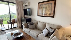 Una sala de estar moderna con un sofá, una mesa de centro, un televisor y un amplio ventanal con vistas a zonas verdes.