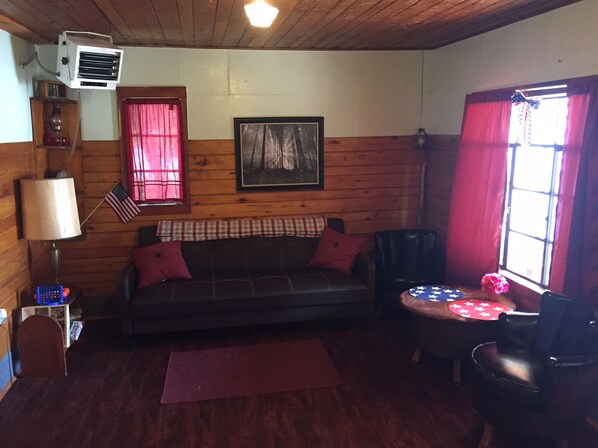 A cozy cabin interior with a wooden floor, a leather sofa, a small table with a red and white tablecloth, and a window with red curtains.