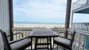 A balcony with a table and chairs, overlooking a beach with people and umbrellas.