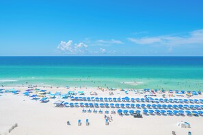 A beach with rows of blue umbrellas and sun loungers, a clear sky, and the ocean in the background.