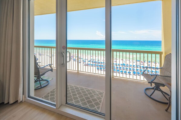 A room with a sliding glass door leading to a balcony with a view of a beach and ocean.