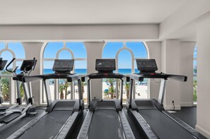 A gym with treadmills and exercise equipment, large windows with a view of the beach and palm trees.