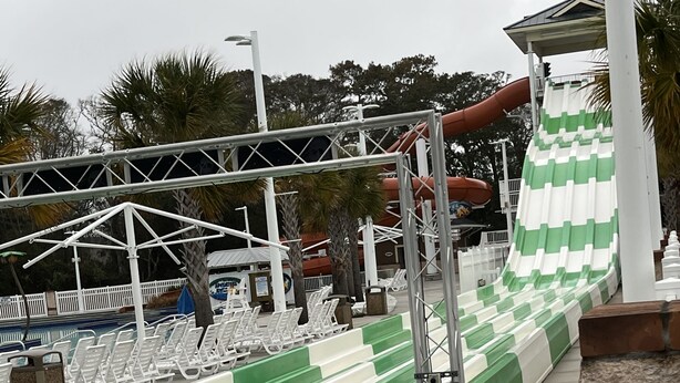 Een waterglijbaan met groene en witte strepen, een overdekt zitgedeelte met witte stoelen en een zwembad met een houten terras.