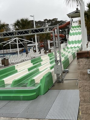 A water slide with green and white stripes, a covered seating area with white chairs, and a pool with a wooden deck.
