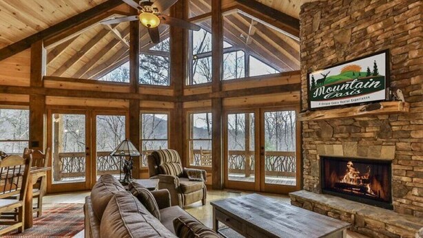 A spacious living room with a stone fireplace, large windows, and a high ceiling. - Golden Oaks at Sugar Creek by Mountain Oasis | Luxury Blue Ridge Cabin Near Ellijay (Blue Ridge)